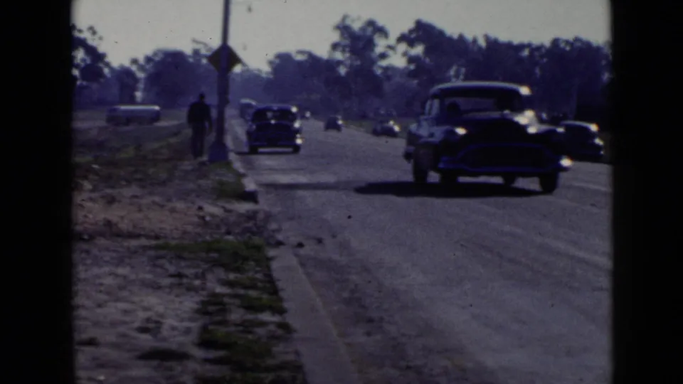 1949 San Diego street view with mid-century cars passing and a dark-clothed pedestrian walking beside the curb.