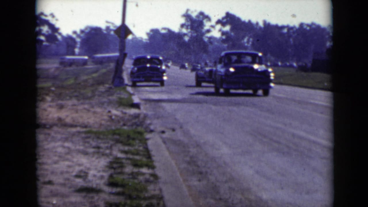 Late-1940s cars move along a sunlit San Diego suburban road, with trees and roadside utility poles in the distance.