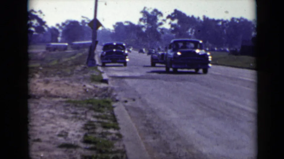Late-1940s cars move along a sunlit San Diego suburban road, with trees and roadside utility poles in the distance.