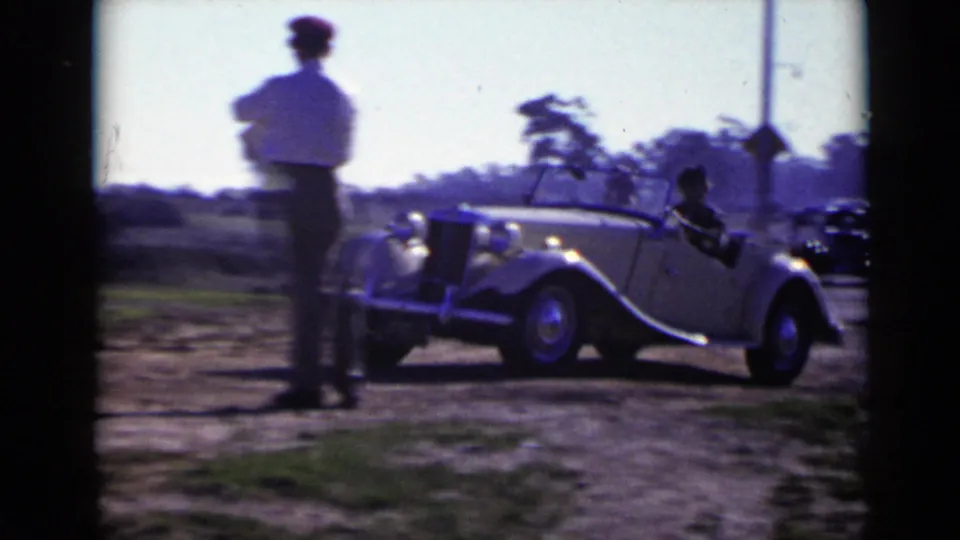 An adult man in a light shirt stands near a parked convertible on a roadside, with trees and a road sign behind him.