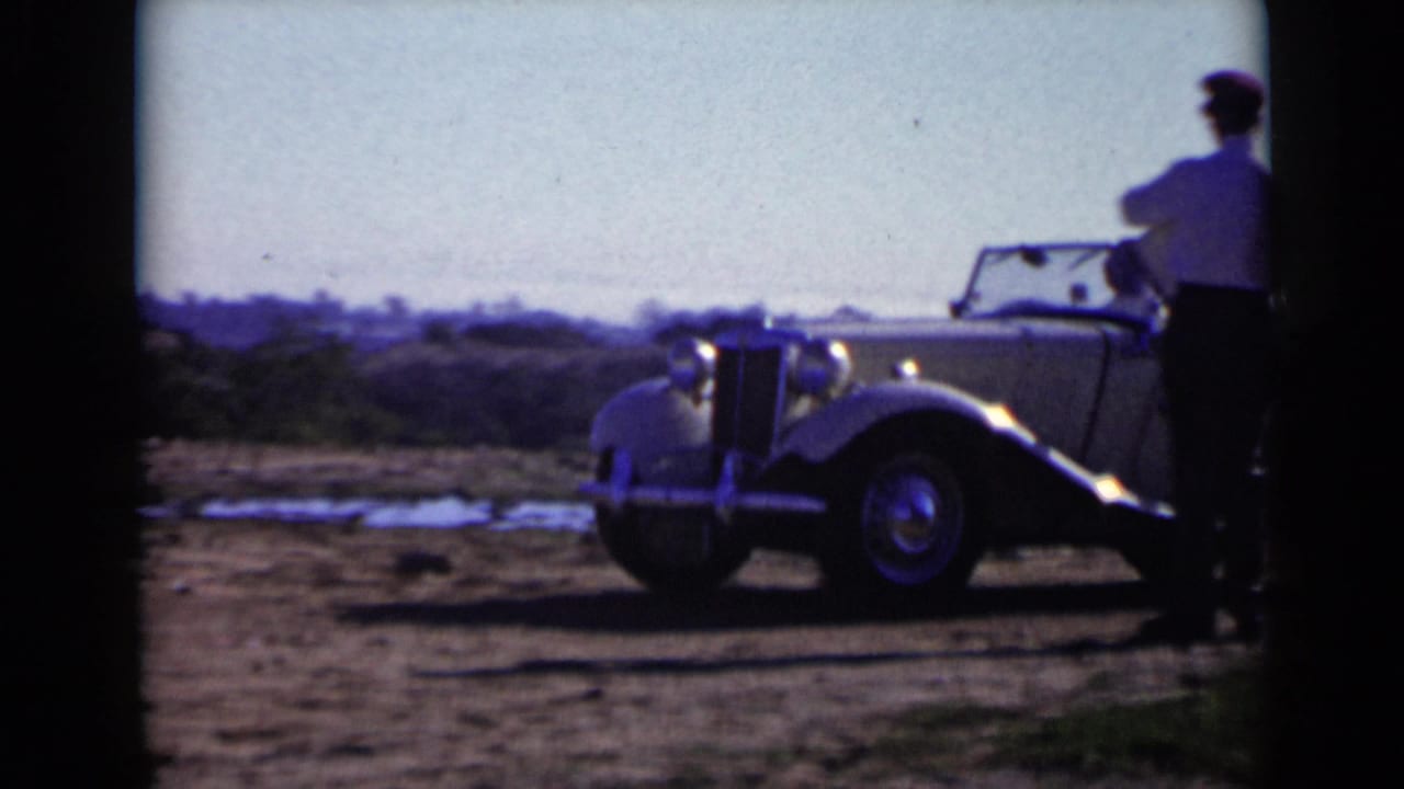 Light-colored antique convertible roadster on a dirt field with a man standing beside it.