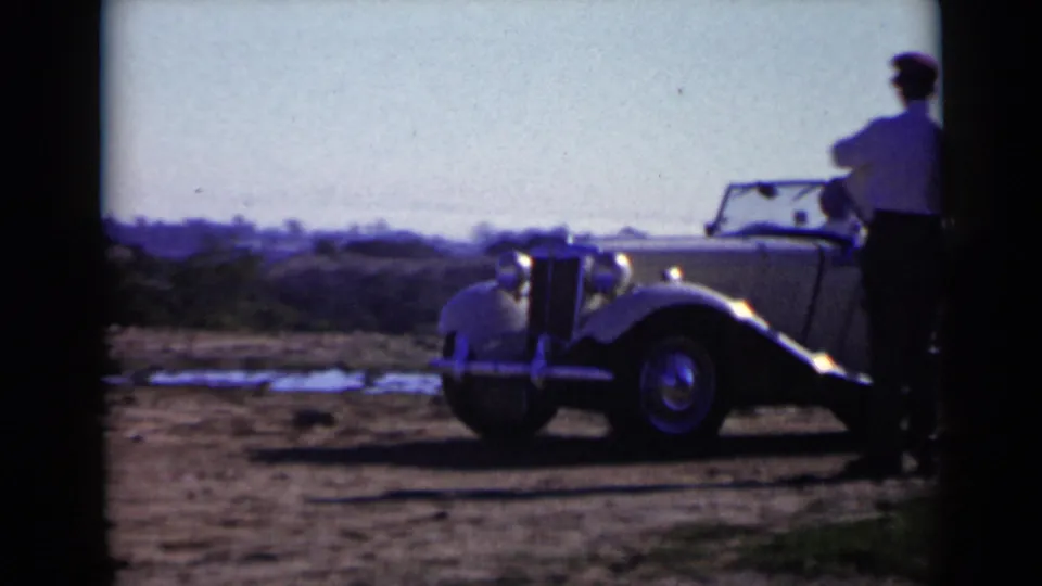 1949 San Diego home-movie frame showing a light-colored antique convertible roadster on open dirt ground with a man nearby.