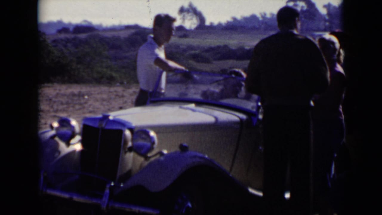 1949 San Diego frame with adults standing around a light-colored antique roadster, its windshield and front grille visible.