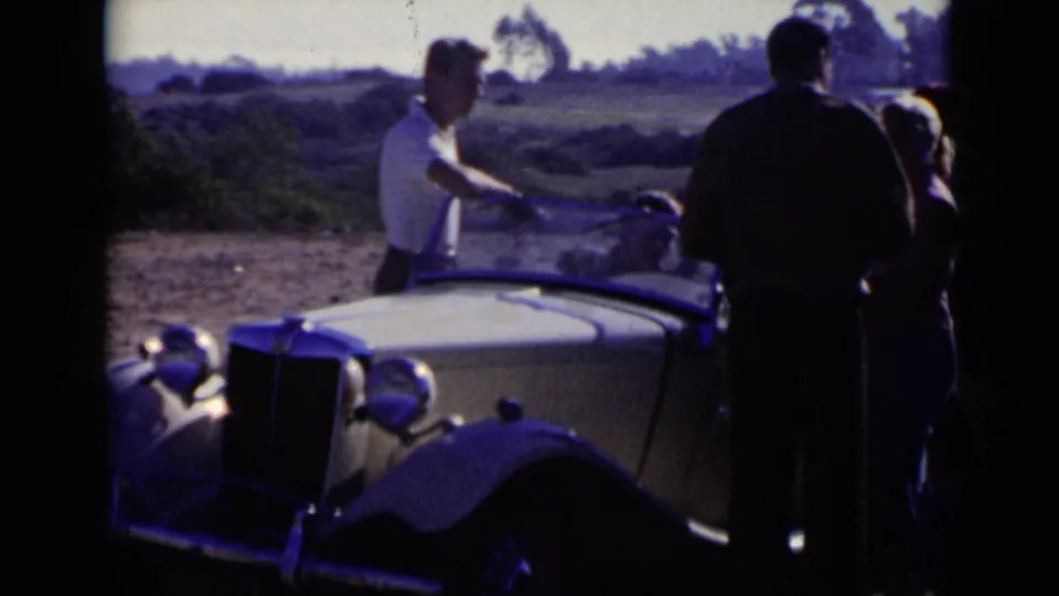 1949 San Diego frame with adults standing around a light-colored antique roadster, its windshield and front grille visible.