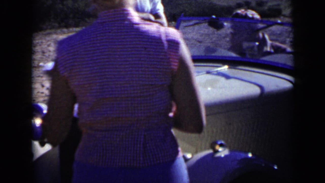 1949 San Diego home-movie frame of a woman from behind near a light open-top car, with other spectators and a dirt road beyond.