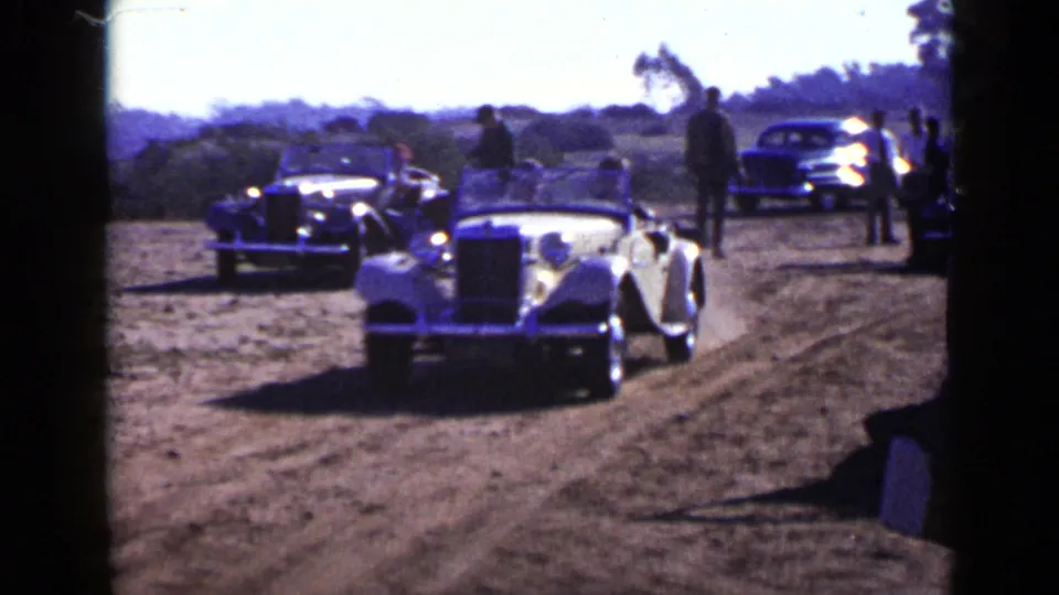 Wide 1949 San Diego frame showing a light convertible among darker cars and spectators spread along a dusty road.