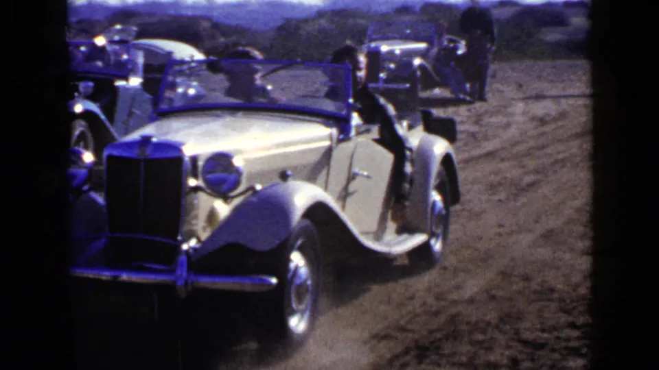 1949 San Diego frame of a cream convertible moving slowly past people and parked cars on a sunlit dirt road.