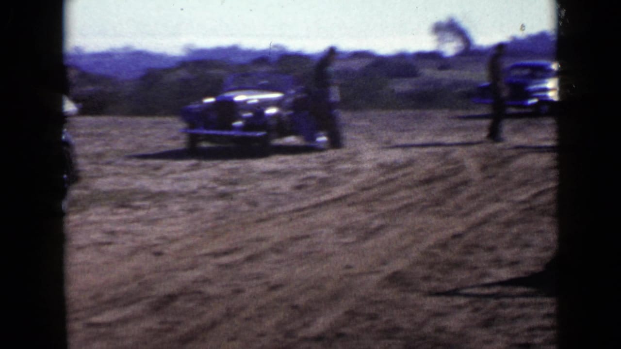 Dark vintage car crossing a dirt road while people stand nearby.