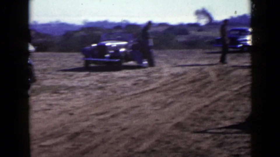 1949 San Diego home-movie frame with a dark vintage car on a dirt turnout and people standing near other automobiles in the background.