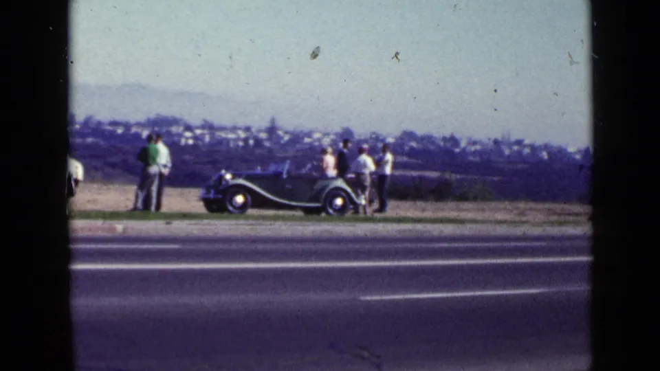 1949 San Diego frame showing adults near a dark roadster beside a paved road, with a distant hillside or city view beyond.