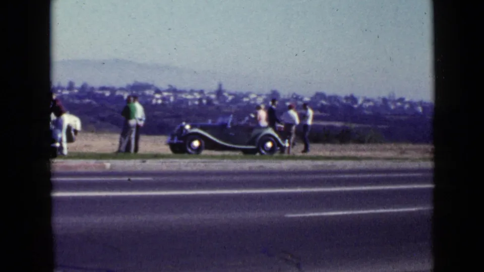 1949 San Diego home-movie frame of people gathered near a dark convertible at the roadside, with a paved road in front and distant landscape behind.