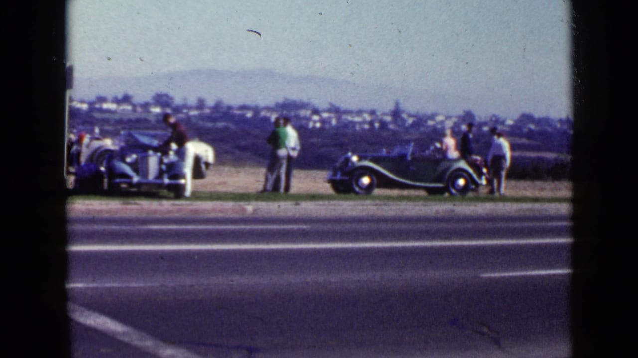 People stand around two classic cars parked off a paved road.