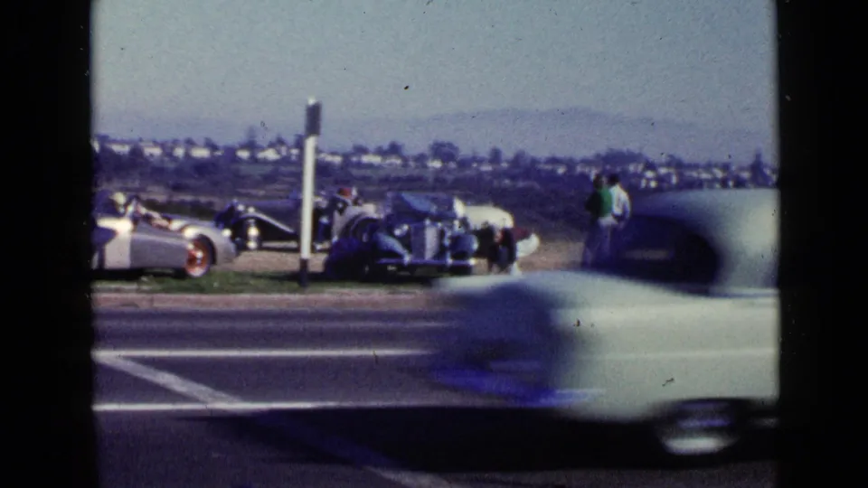 San Diego roadside in 1949 with a blurred passing car in front of adults gathered by classic cars.