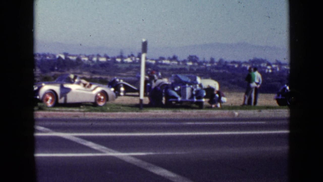 Silver convertible and dark car parked beside a road with adults standing nearby.