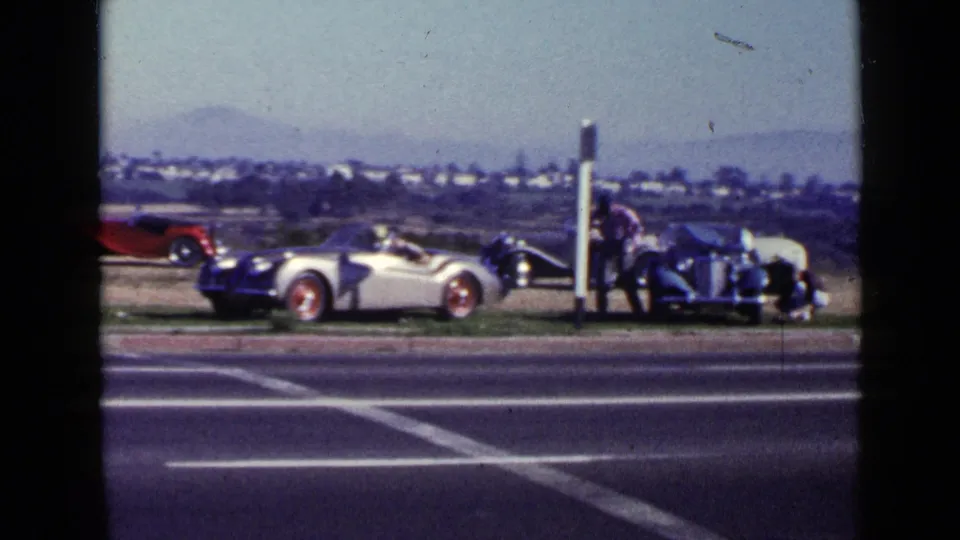 1949 San Diego roadside view with a silver convertible, red roadster, dark vintage car, and an adult walking near the vehicles.