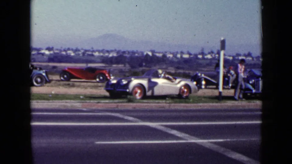 1949 San Diego home-movie frame of a roadside car gathering with a red roadster, silver convertible, blue car, and walking adult.