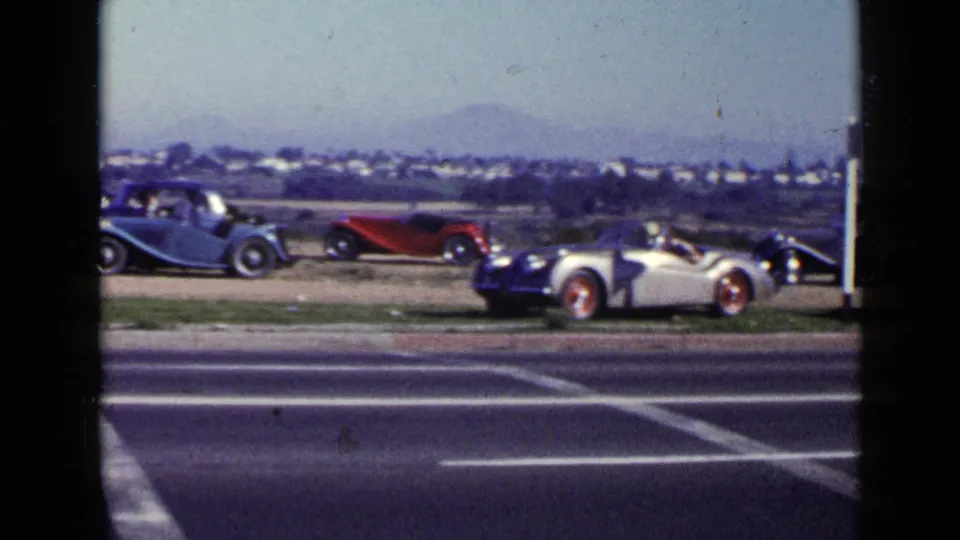 1949 San Diego frame showing multiple vintage automobiles parked along a road, with houses and low hills in the background.