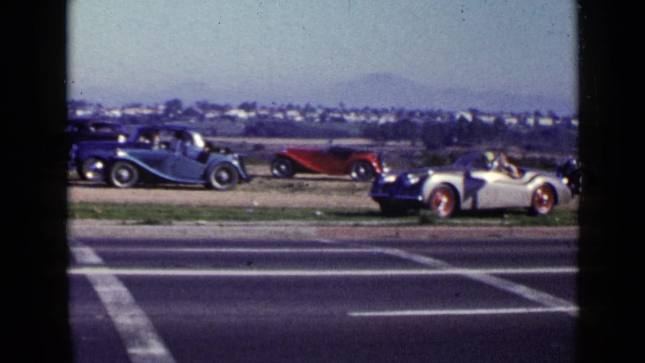 1949 San Diego home-movie frame showing parked vintage roadsters beside a roadway with hills and a low skyline beyond.