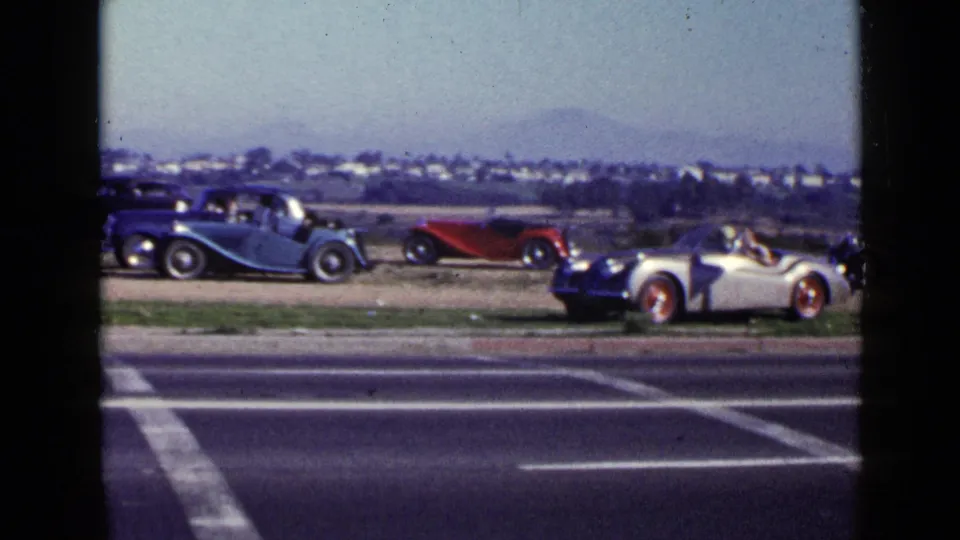 1949 San Diego home-movie frame showing parked vintage roadsters beside a roadway with hills and a low skyline beyond.