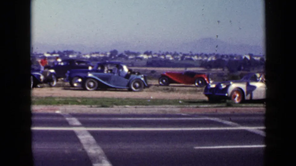 San Diego 1949 footage frame with classic cars parked beyond a road and a pale sports car partly visible at the right.