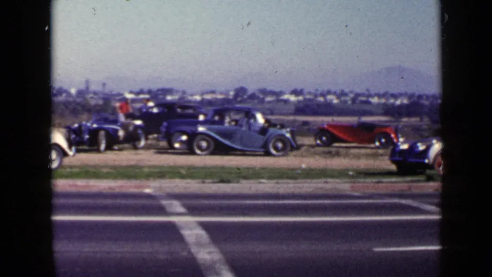 1949 San Diego home-movie frame showing a roadside lineup of vintage cars and a small person in red near the display.