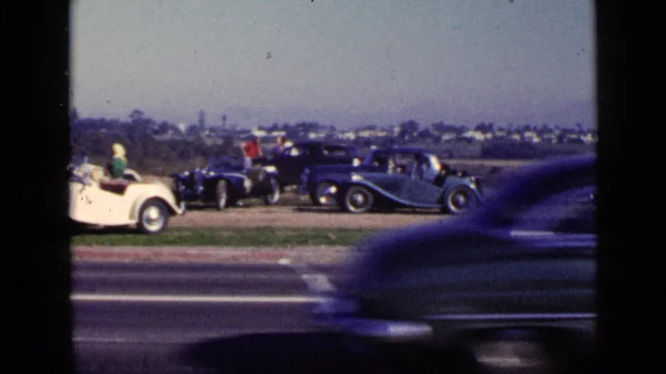 San Diego 1949 frame with classic cars and spectators beyond the road, partly obscured by a passing foreground vehicle.