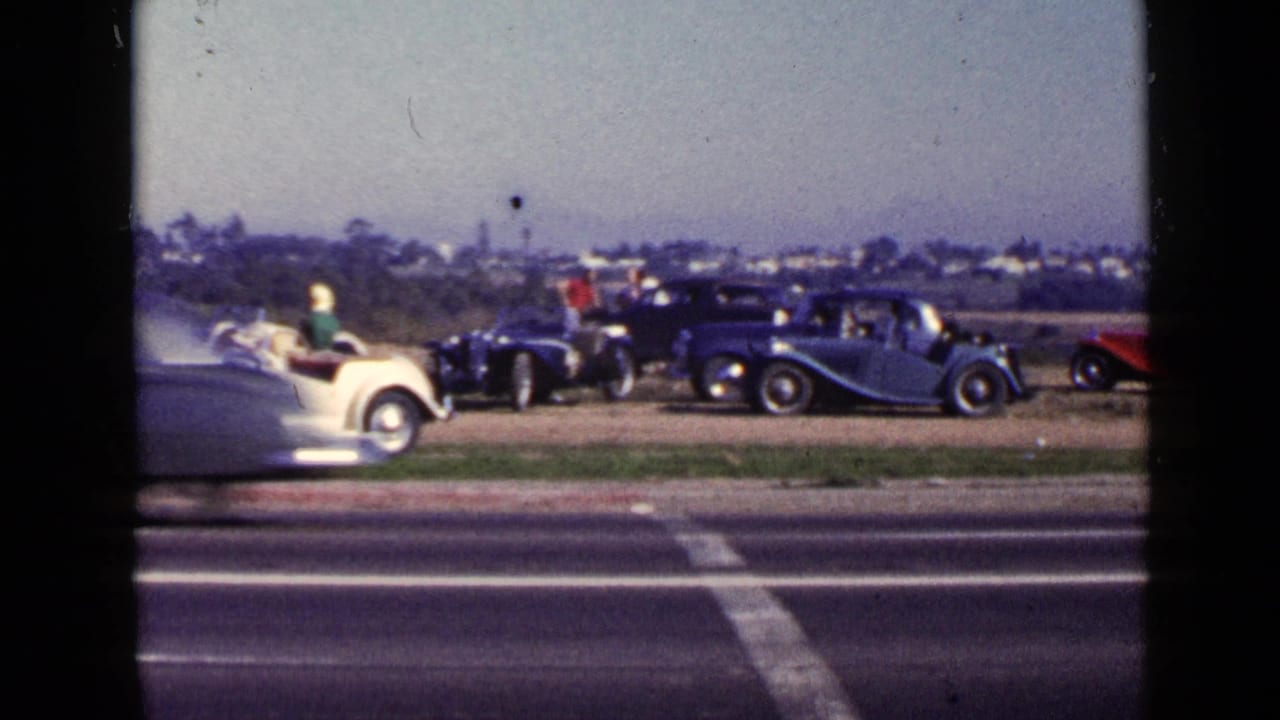 White convertible and dark classic cars parked beyond a roadside with people nearby.