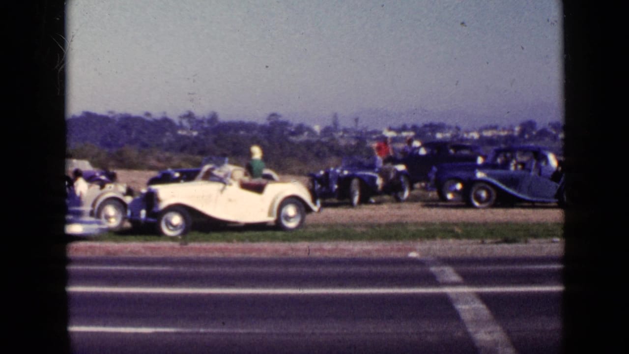 White convertible in front of several dark classic cars beside a road.