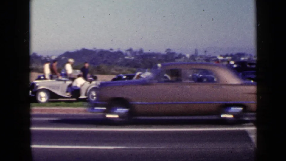 A passing sedan creates motion blur in front of people and parked classic cars along a San Diego roadside in 1949.