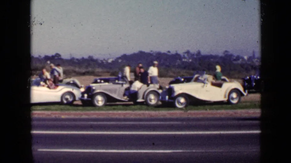 Groups of people stand and sit near white and dark classic cars parked along a grassy roadside in 1949 San Diego.