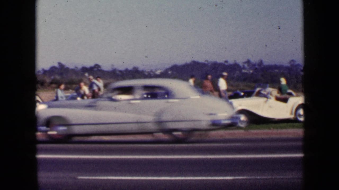 1949 San Diego home-movie frame showing adults near parked classic cars while a light-colored sedan blurs across the road.