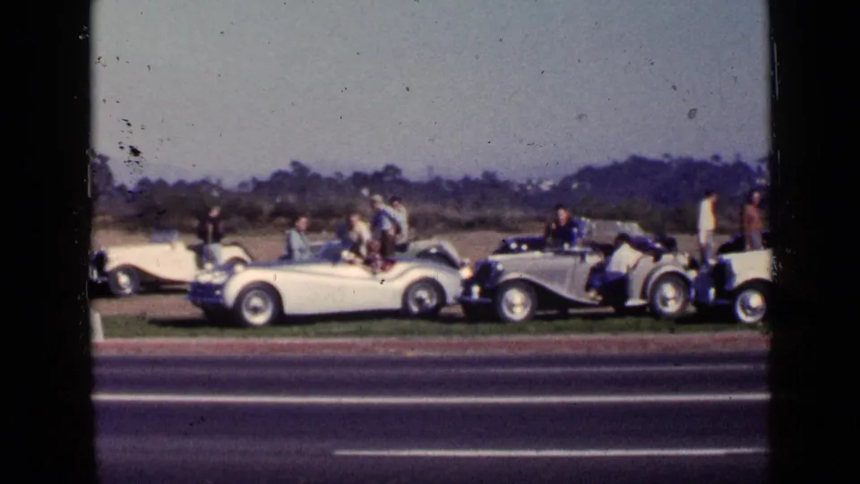 1949 San Diego frame with people standing and sitting near white and silver convertibles on a grassy roadside verge.