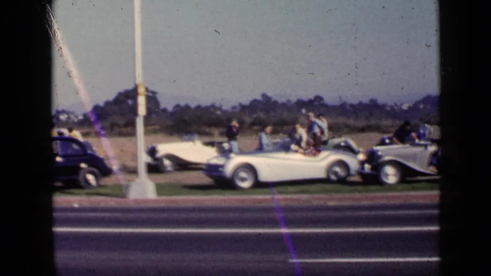 1949 San Diego home-movie frame showing parked roadsters, adults gathered beside them, and a roadside pole near the curb.