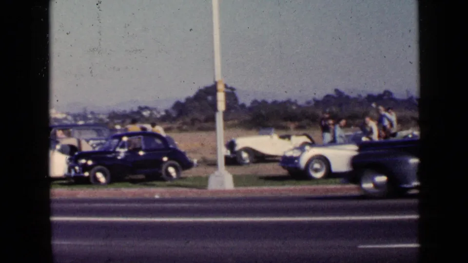 1949 San Diego frame with a dark sedan moving past adults and classic cars parked beside the highway.