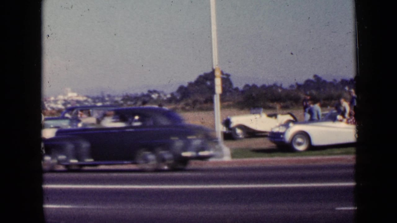 San Diego 1949 home-movie frame with a dark sedan blurred in the foreground and parked classic cars with adults beyond the road.