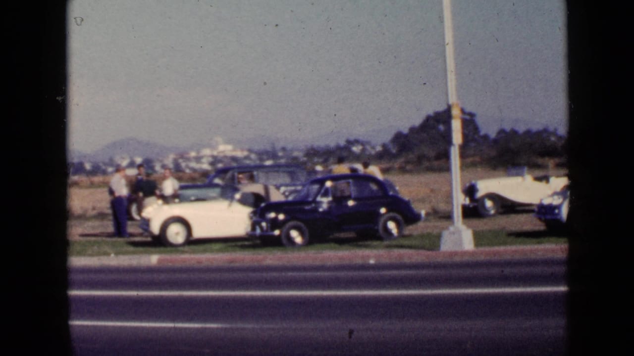 San Diego 1949 roadside car scene showing adults gathered near parked white and dark automobiles beyond a multilane road.