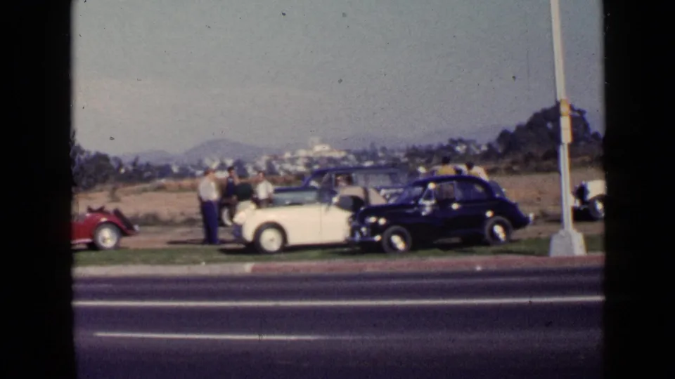 San Diego 1949 frame with adults beside white and dark parked cars, a red roadster at the edge, and a highway in front.