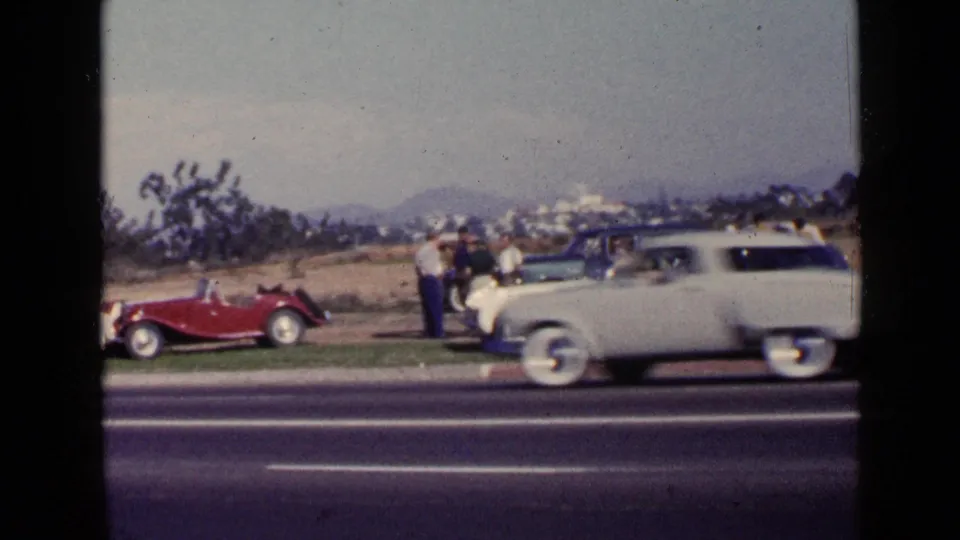 San Diego 1949 home-movie frame with a light sedan passing in front of adults and parked classic cars on a grassy verge.