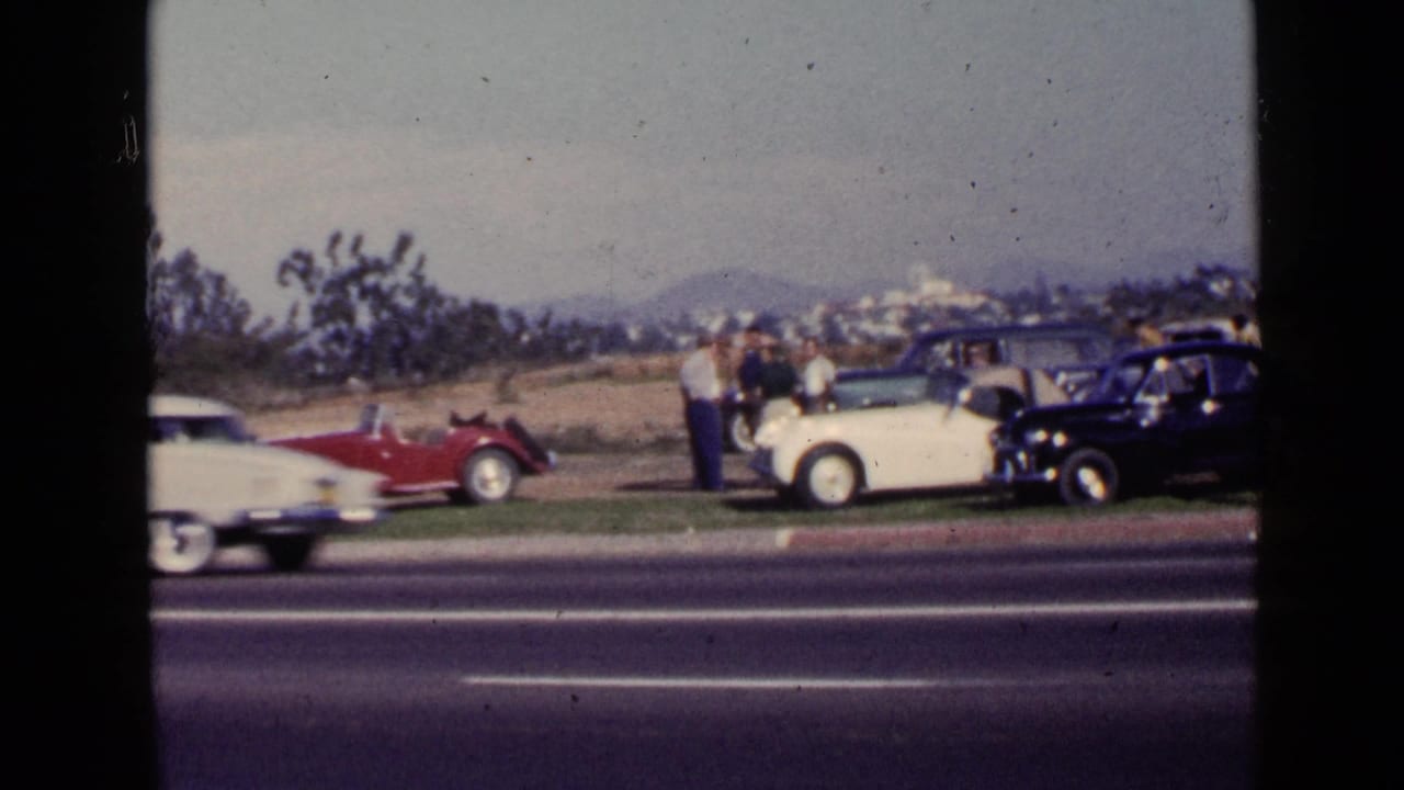 1949 San Diego home-movie frame showing antique cars on a grassy verge beside a highway with adults gathered nearby.