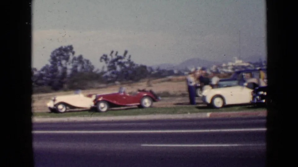 1949 San Diego frame with red and cream antique cars parked beside a highway as adults gather at the right edge.