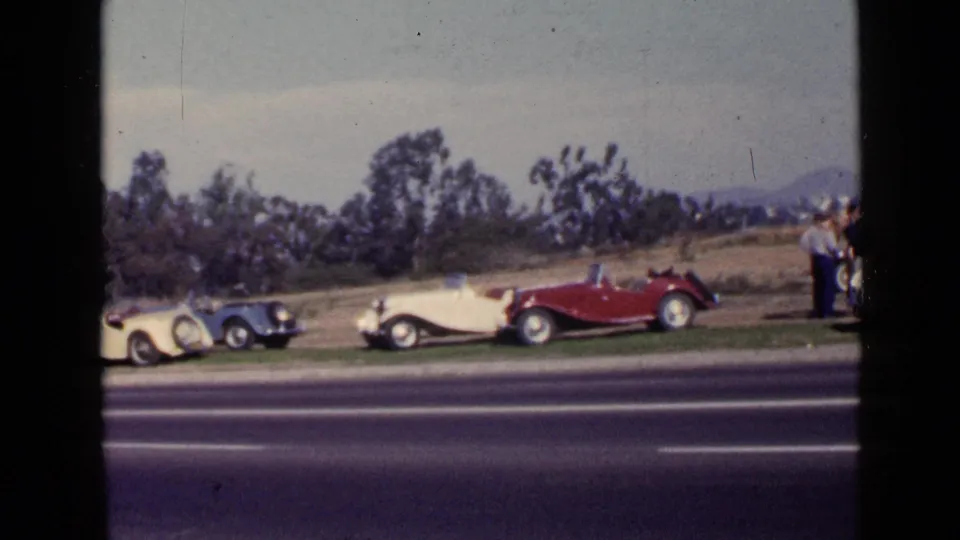 1949 San Diego frame showing a row of antique cars beside a highway, with adults partly visible near the right edge.