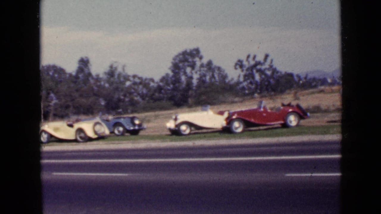 San Diego 1949 home-movie frame showing cream, blue, and red open-top cars lined up beside a road with trees and hills beyond.
