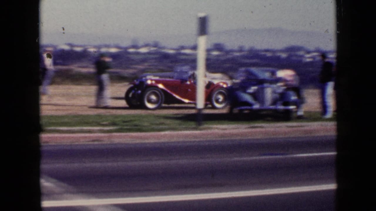 San Diego 1949 frame with a red open-top roadster, a blue vintage car, and several spectators on the roadside verge.