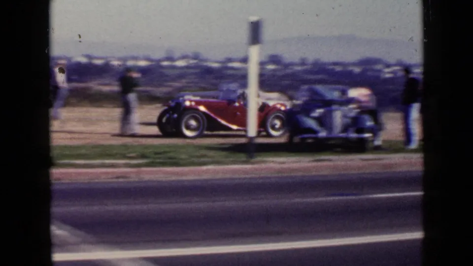 San Diego 1949 frame with a red open-top roadster, a blue vintage car, and several spectators on the roadside verge.