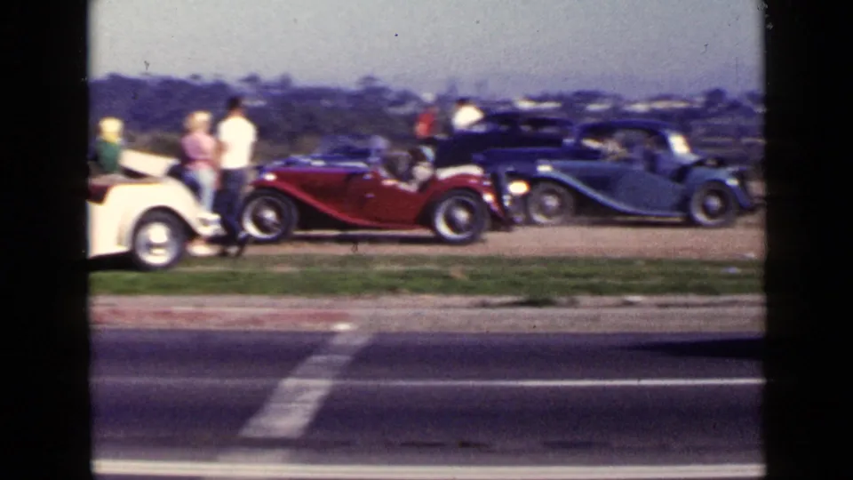 San Diego 1949 roadside scene with spectators gathered around cream, red, and blue open-top cars on a grassy verge.