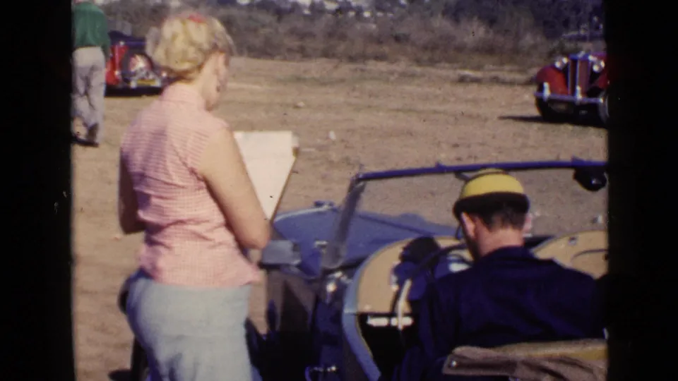San Diego 1949 home-movie frame showing a woman in a pink checkered top and a man in a yellow cap beside a blue open car.