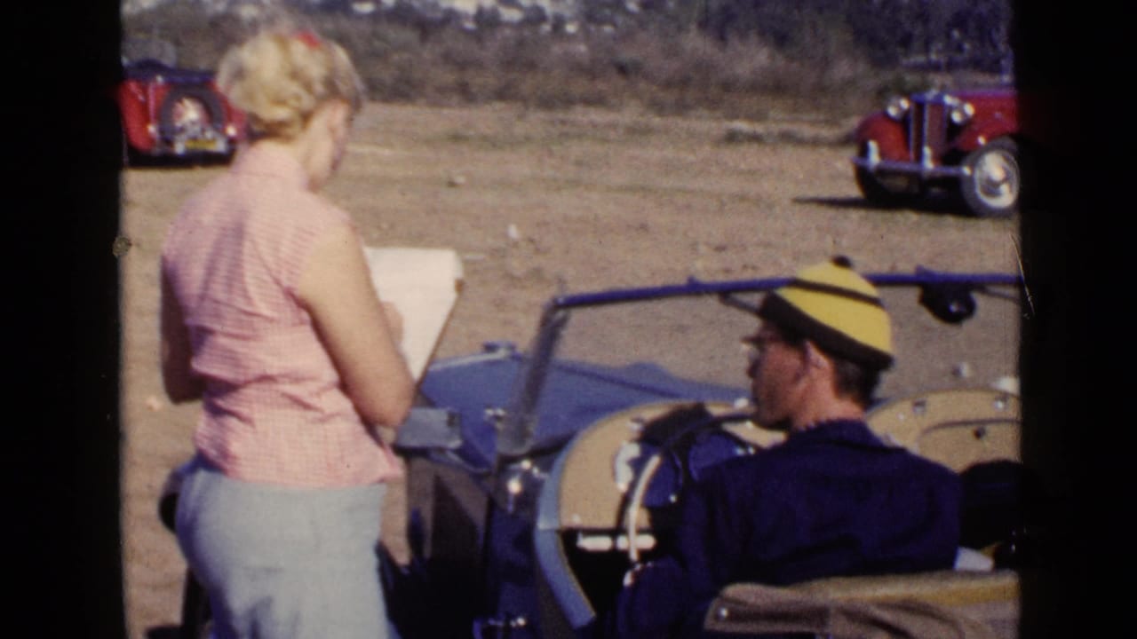 Woman holding papers beside a blue convertible with a seated driver in a striped cap.