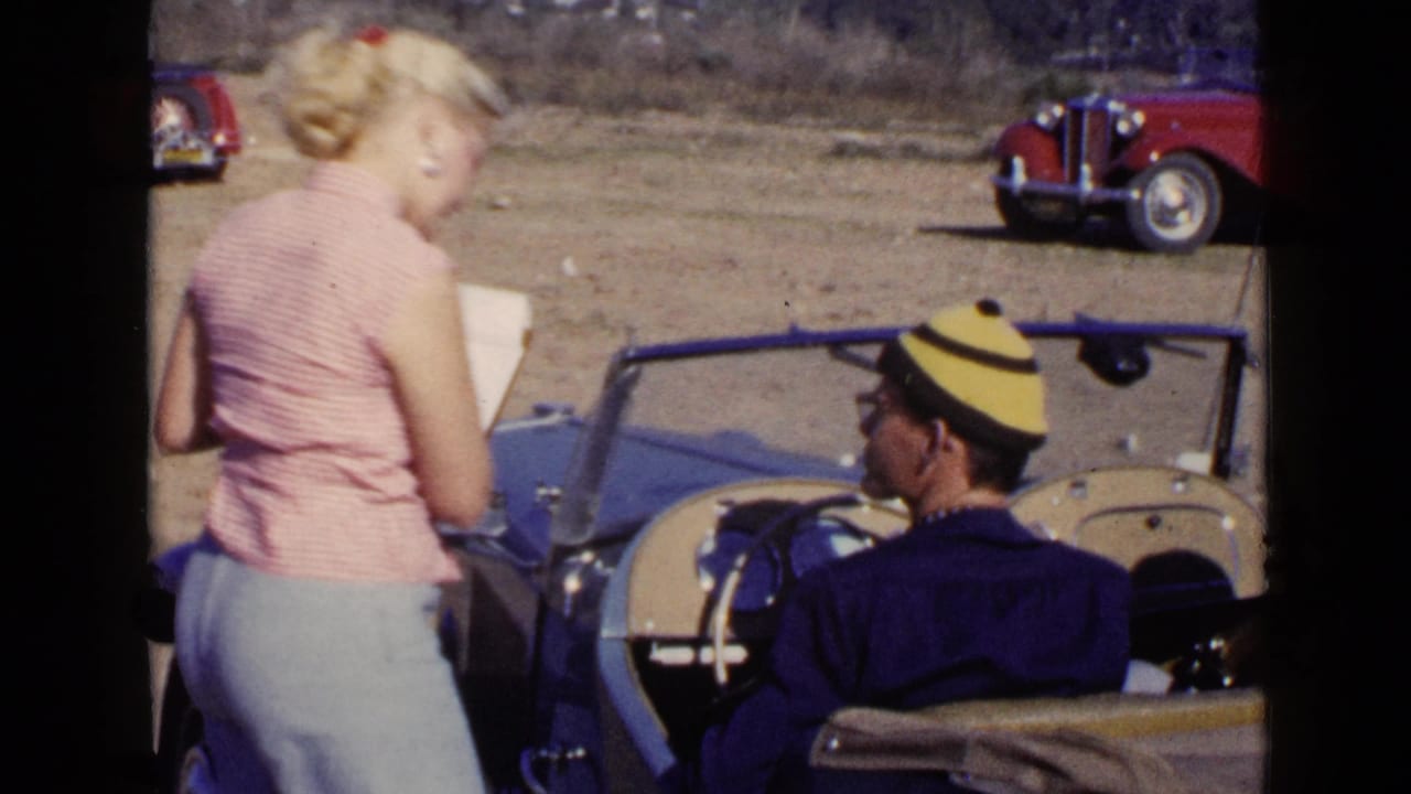 Woman turning with papers near a blue convertible while the driver sits inside.