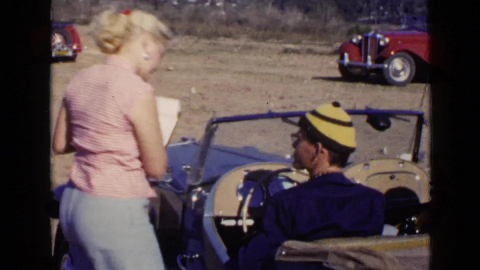 The woman turns slightly with paperwork next to the blue convertible, with the driver seated and a red car visible behind them.