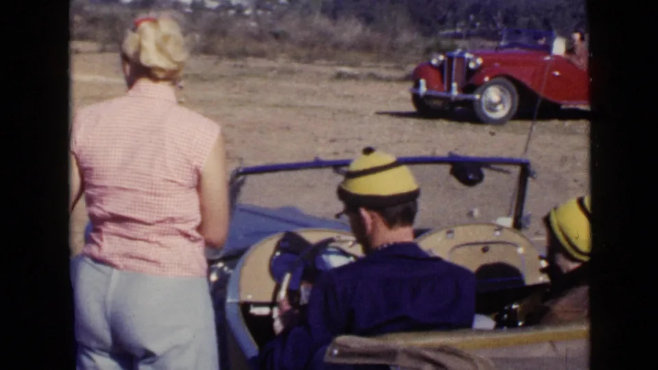 A rear view shows the woman by the blue convertible, the driver seated below the windshield, and a red vintage car in the field.